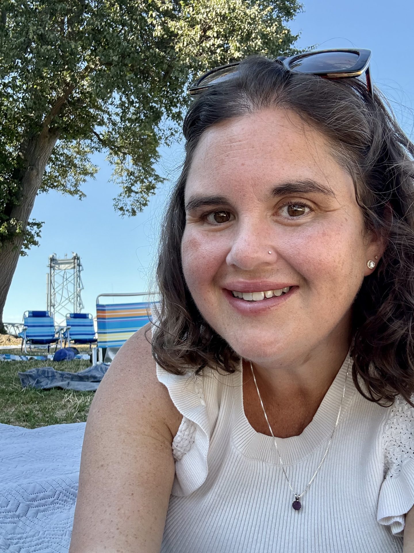 A smiling woman with shoulder-length wavy dark hair lies on a blanket outdoors in a grassy park. She wears a light-colored sleeveless top, a necklace with a small purple pendant, and sunglasses resting on her head. Behind her are folding beach chairs, a large leafy tree, and a tall metal structure against a bright blue sky.