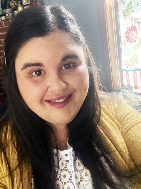 A woman with long brown hair , brown eyes and a smile - wearing a yellow cardigan and white top